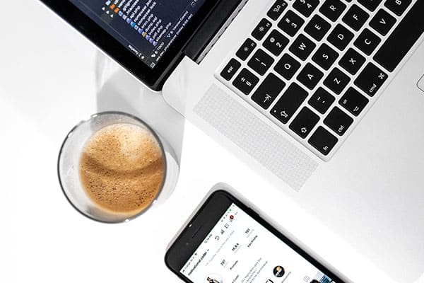 A close-up view of a laptop keyboard, a glass of coffee, and a smartphone displaying a social media app on a white surface.
