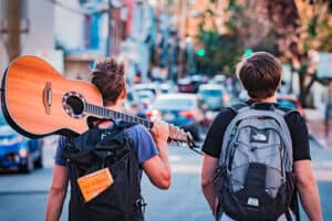 Two people with backpacks walk down a city street. One carries an acoustic guitar on their shoulder, and a book titled The 4-Hour Workweek sticks out of their backpack. Cars and buildings line the street under a partly cloudy sky.