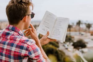 A person wearing sunglasses and a plaid shirt reads and contemplates a book with diagrams while sitting outdoors overlooking a blurry, sunlit cityscape.