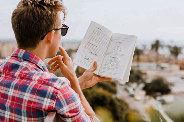 A person wearing sunglasses and a plaid shirt reads and contemplates a book with diagrams while sitting outdoors overlooking a blurry, sunlit cityscape.