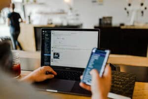 Person sits at a wooden table using a laptop with a messaging app open, while holding a smartphone in their other hand. A drink sits to the left of the laptop and another person is seen blurred in the background.