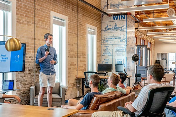 A man stands and gives a presentation to four seated people in a modern, open office space with brick walls, large windows, and visible brainstorming diagrams on one wall.