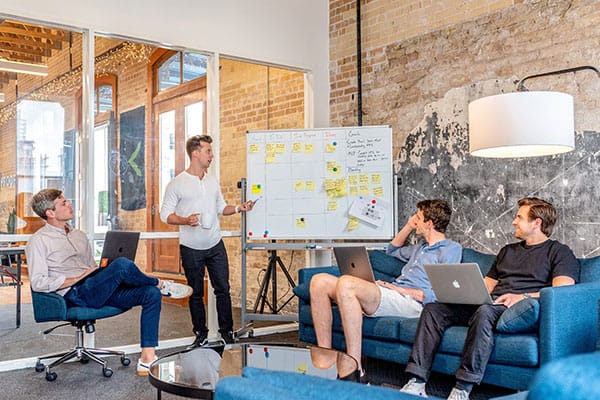 Four people in a modern office setting, three seated with laptops and one standing, present and discuss ideas at a whiteboard covered with sticky notes and writing.