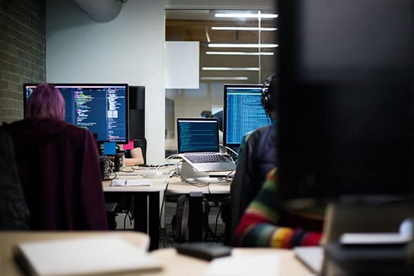 Two people sit at desks with multiple computer monitors displaying code in an office setting. Cables, notebooks, and a laptop are visible on the desks. The workspace is modern with glass walls and overhead lighting.