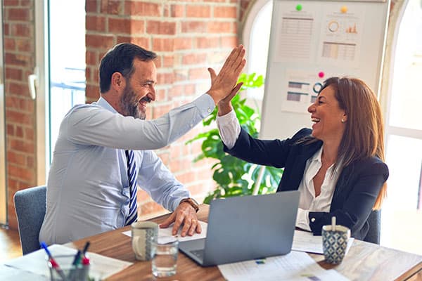 Two colleagues in business attire sit at a desk with papers and a laptop, smiling and giving each other a high five in an office with brick walls and charts on a board in the background.
