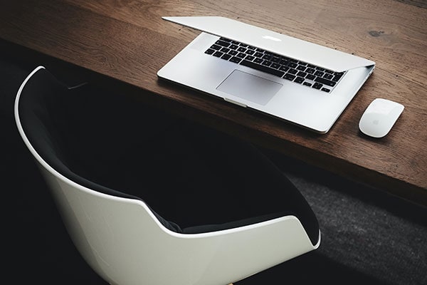 Partially closed laptop and wireless mouse on a wooden desk next to a modern black and white chair, viewed from above.