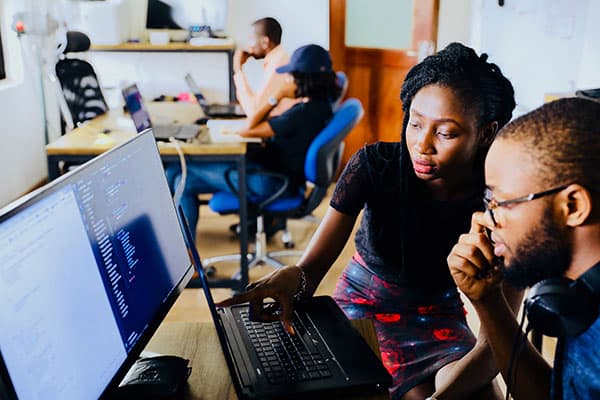 Two people sit at a desk working together on a computer displaying code, while others work at separate desks in the background.