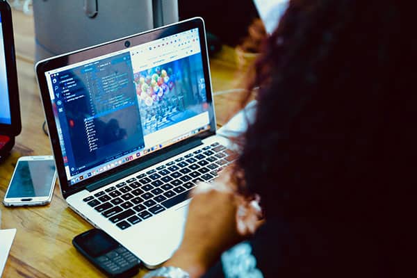 Person working at a desk on a laptop displaying coding software and a web browser. A smartphone and a basic mobile phone are on the desk nearby. The person’s face is not visible.