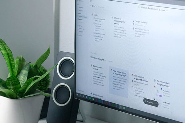 A computer monitor displays a project management or note-taking app with organized sections and tasks. A small green potted plant and black speakers are positioned next to the monitor on a white desk.