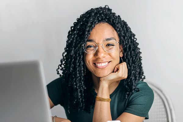 A person with curly hair and glasses, wearing a dark green shirt, sits at a desk with a laptop, smiling at the camera and resting their chin on their hand.