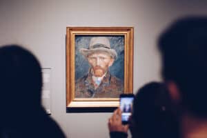 A framed portrait painting of a bearded man wearing a hat hangs on a gallery wall as visitors view and photograph it.