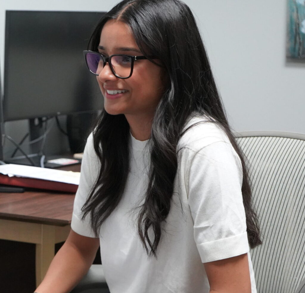 A woman with long dark hair and glasses sits in an office chair, holding a notepad and pen, and smiles at someone off-camera. A desk with a computer monitor is visible in the background.